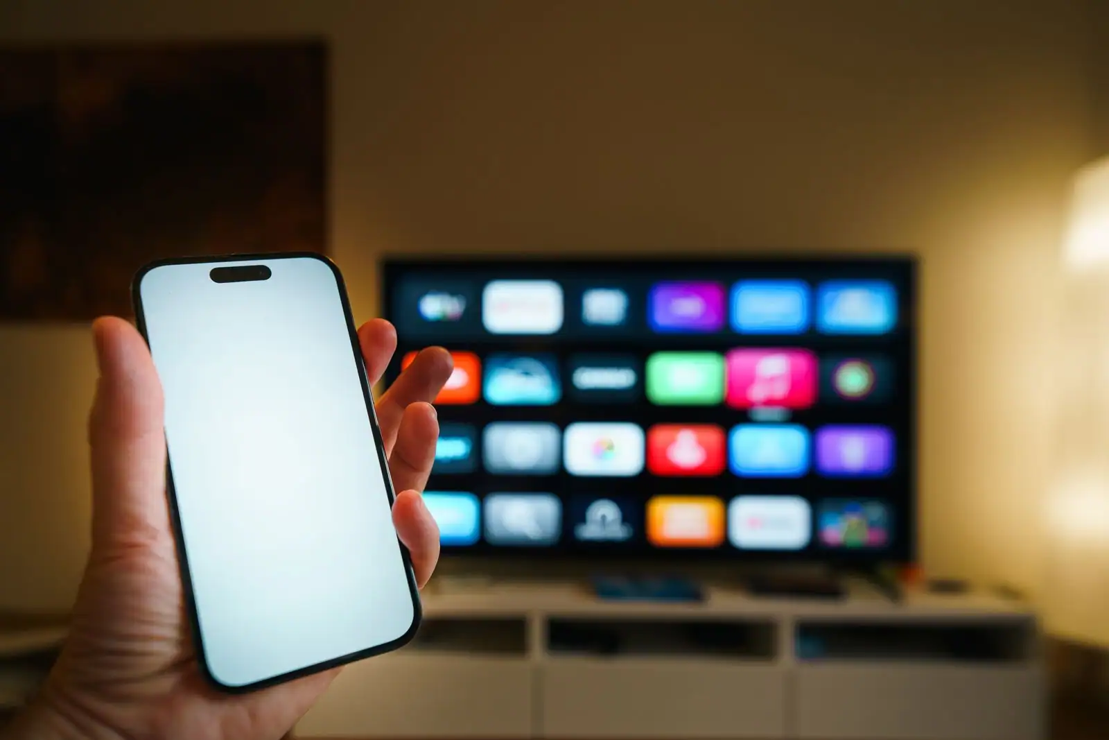Main tenant un smartphone devant un téléviseur connecté affichant une grille d'applications dans un salon