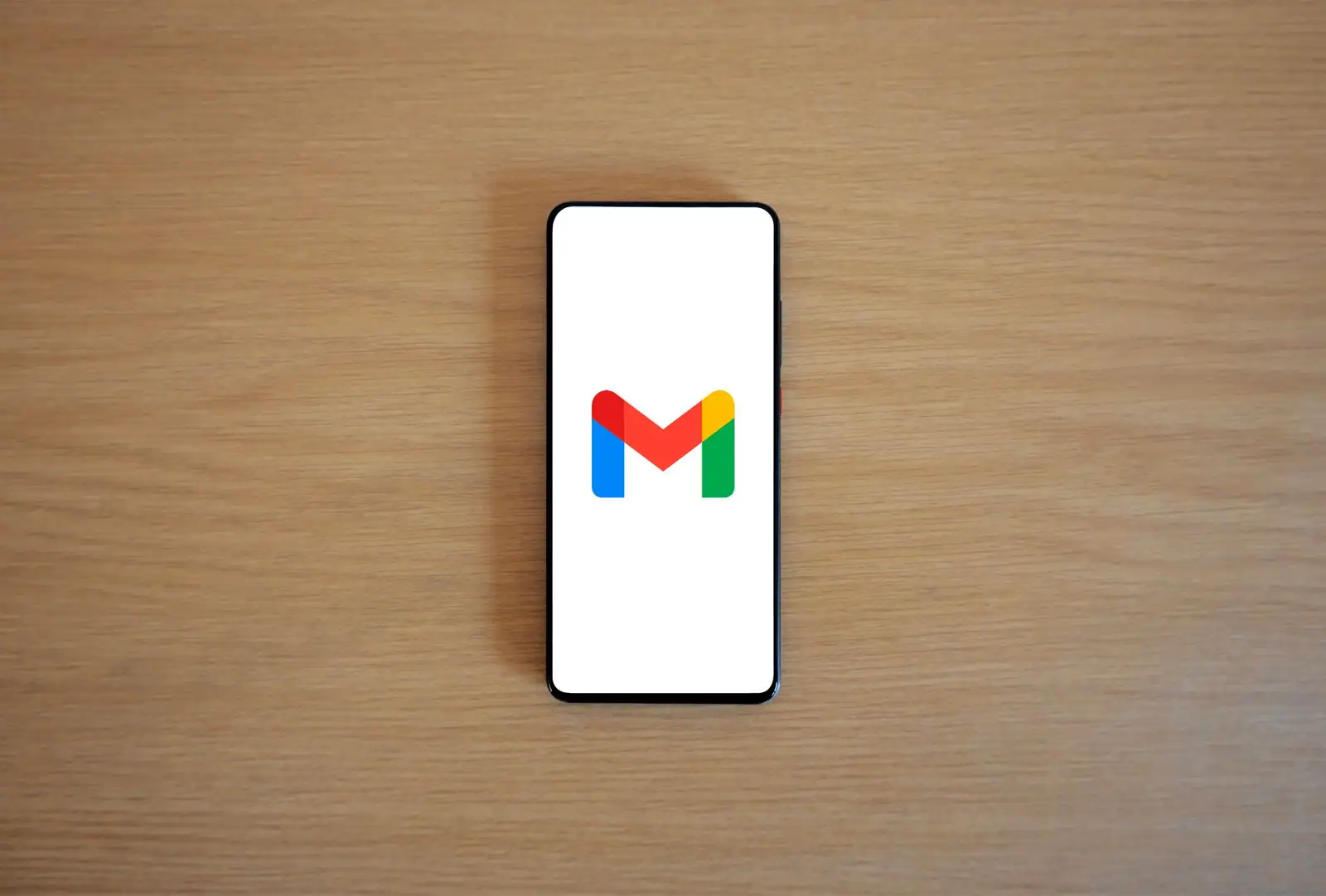 Un smartphone posé sur un bureau en bois affiche le logo coloré de l'application Gmail.