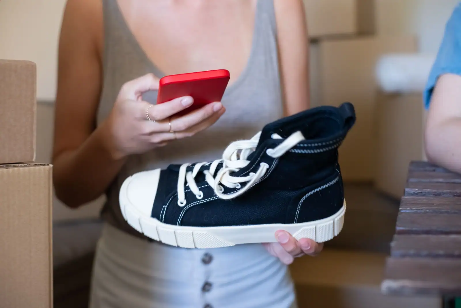 Personne tenant une basket d'occasion et un smartphone près de cartons, prête à la vendre en ligne