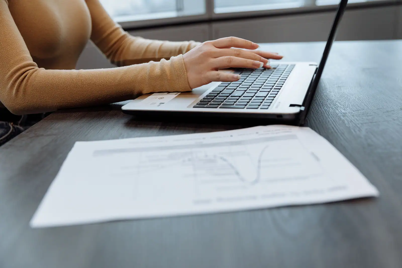 Mains d’une personne sur le clavier d’un ordinateur portable, un document imprimé posé devant elle.
