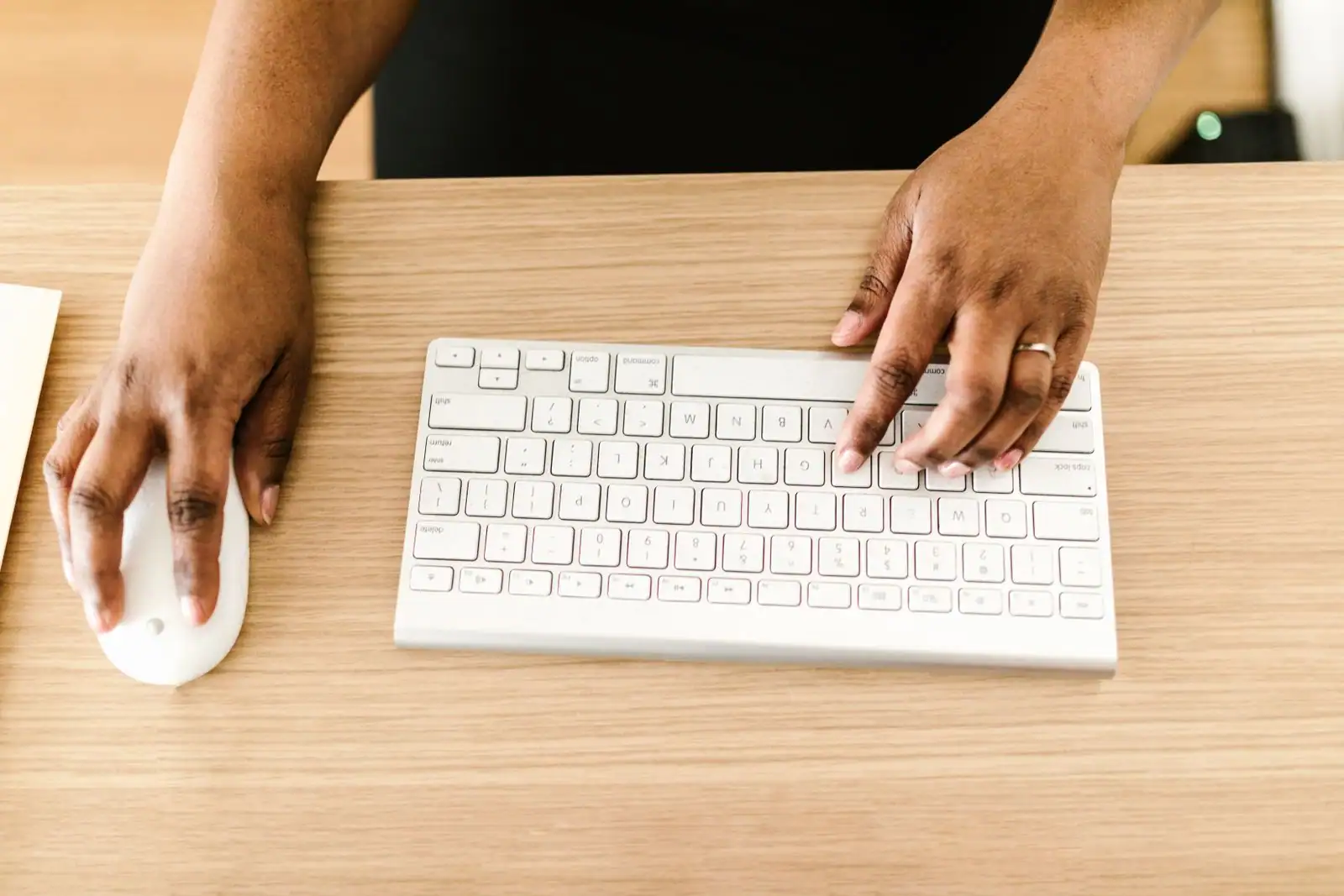 Mains posées sur un clavier d'ordinateur et une souris, sur un bureau en bois clair.