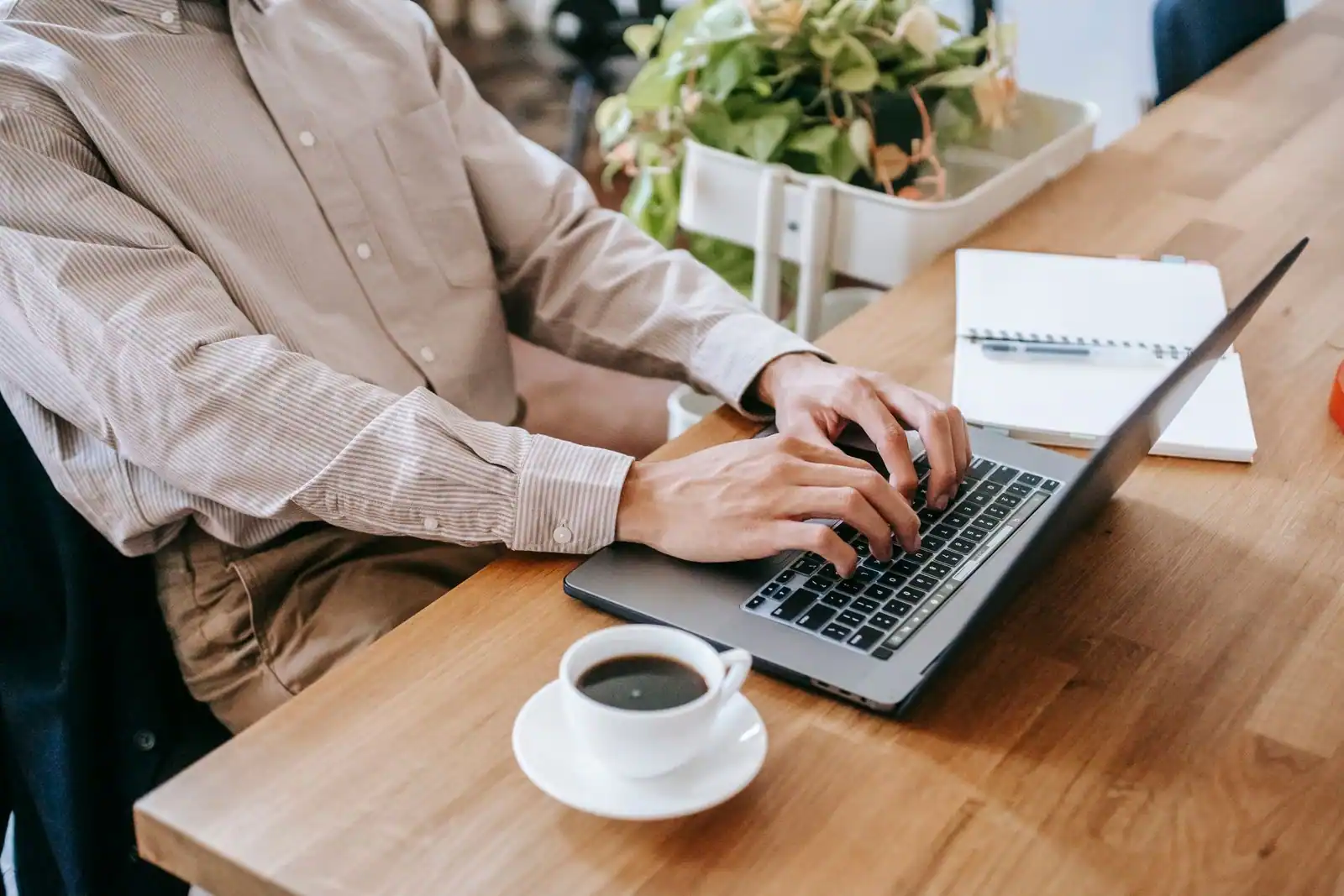 Mains sur le clavier d'un ordinateur portable, carnet et tasse de café sur une table en bois clair