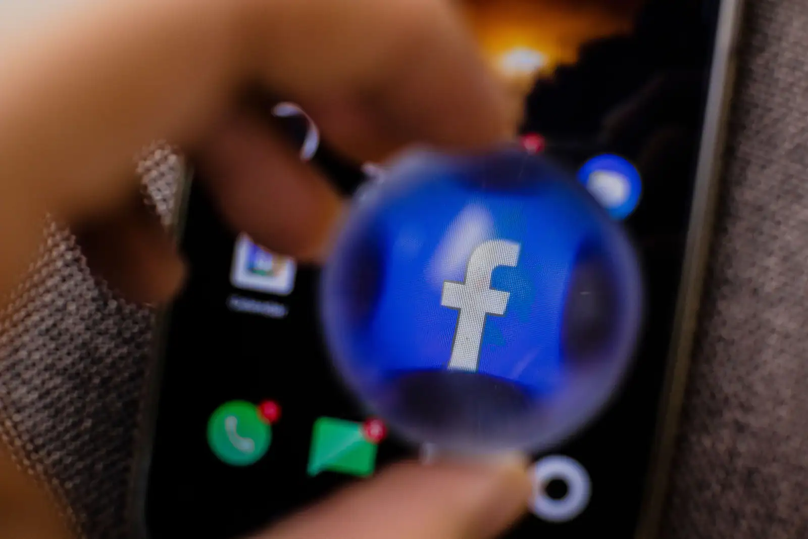 Loupe posée sur l’écran d’un smartphone, grossissant l’icône bleue de l’application Facebook.