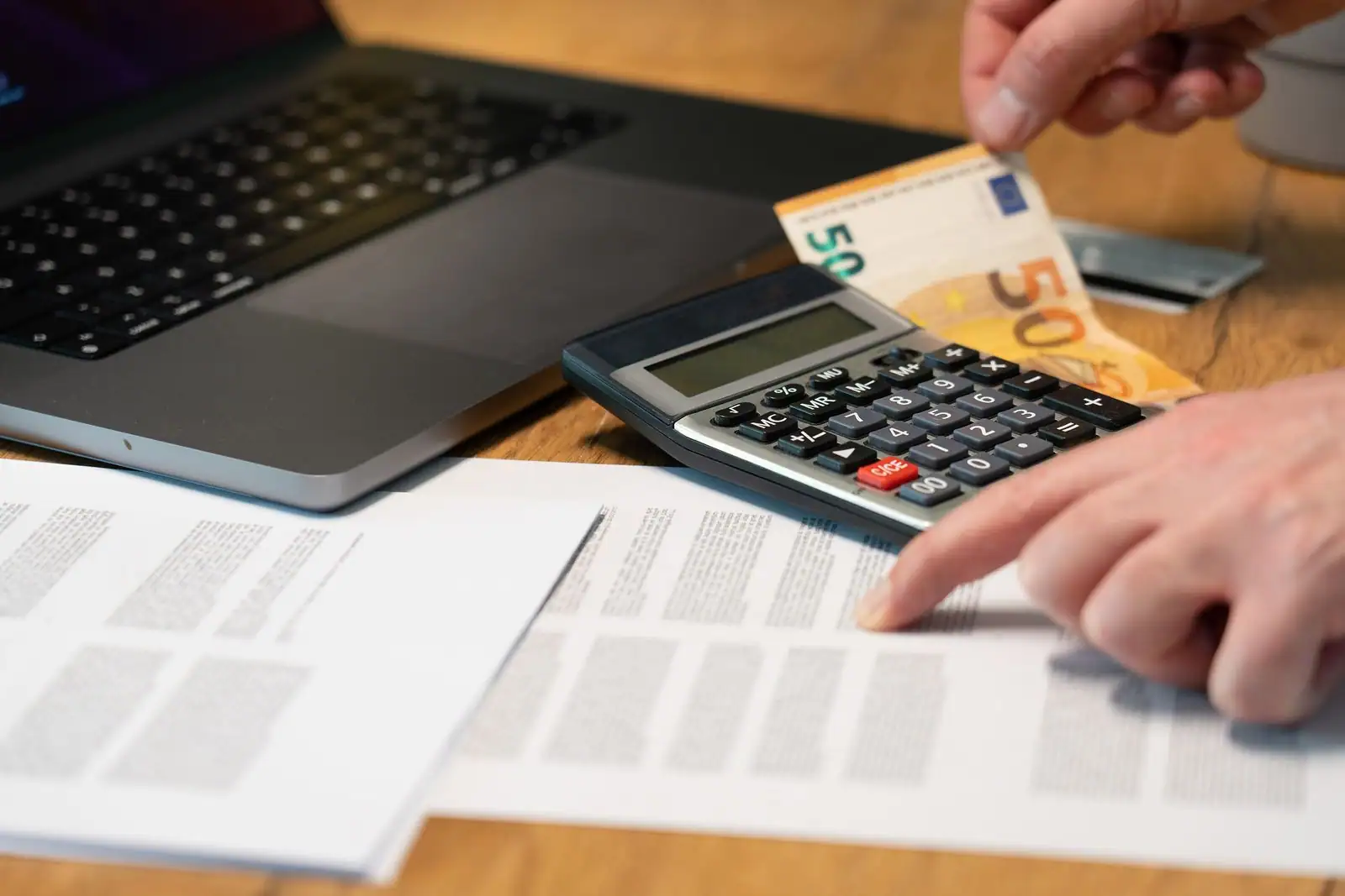 Main tenant un billet de cinquante euros au-dessus d'une calculatrice, documents et ordinateur portable