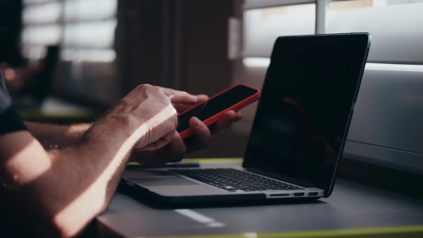 Les mains d'une personne utilisant un téléphone devant un ordinateur portable ouvert, posé sur un bureau.