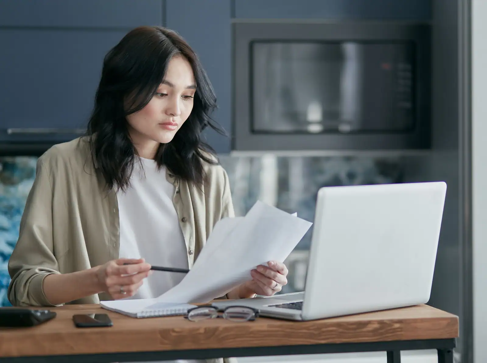 Une femme assise à une table de cuisine relit des documents papier devant un ordinateur portable ouvert.