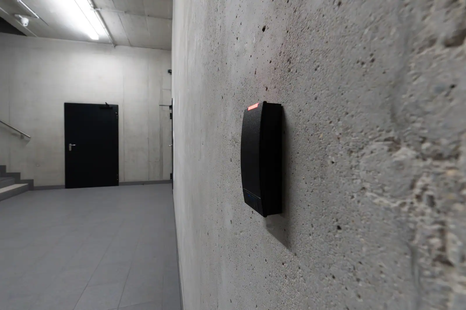 Lecteur de badge noir fixé sur un mur en béton, devant une porte fermée dans un couloir de bureau.