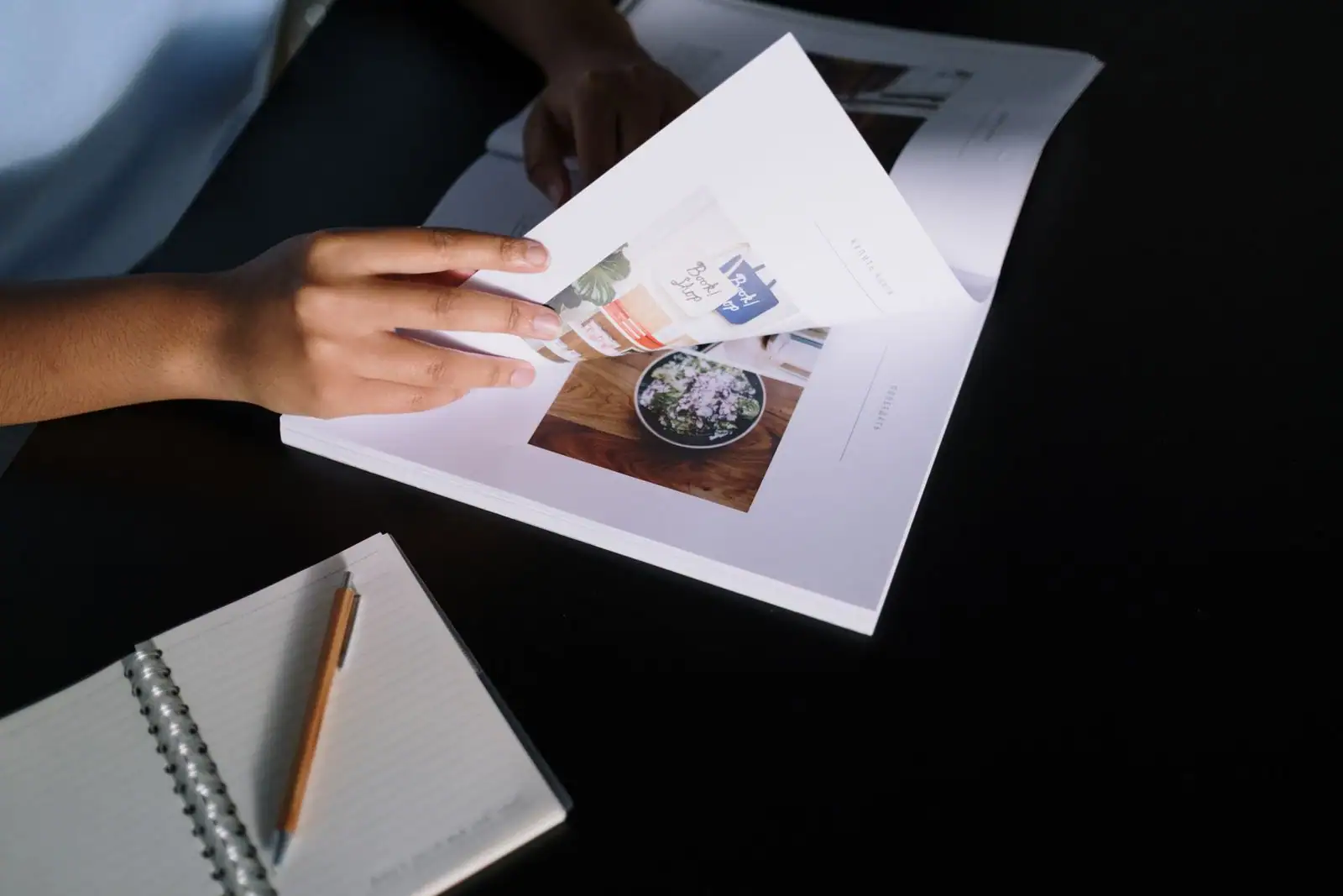 Mains feuilletant les pages d'une revue imprimée posée sur une table sombre, à côté d'un carnet à spirale